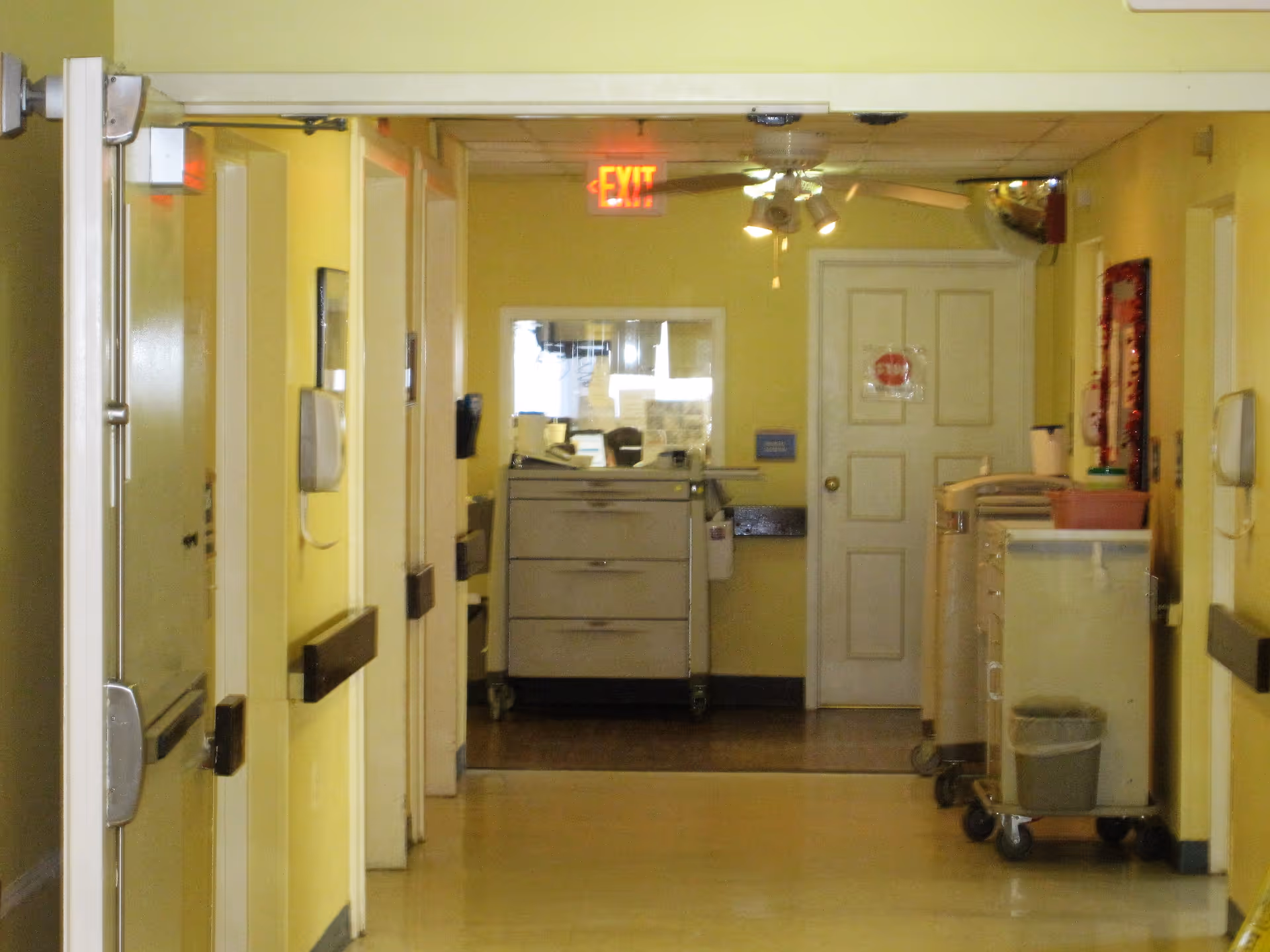 A hallway inside a healthcare facility with yellow walls, several doors on each side, a ceiling fan with lights, an exit sign above a door at the end, and medical carts along the right side.