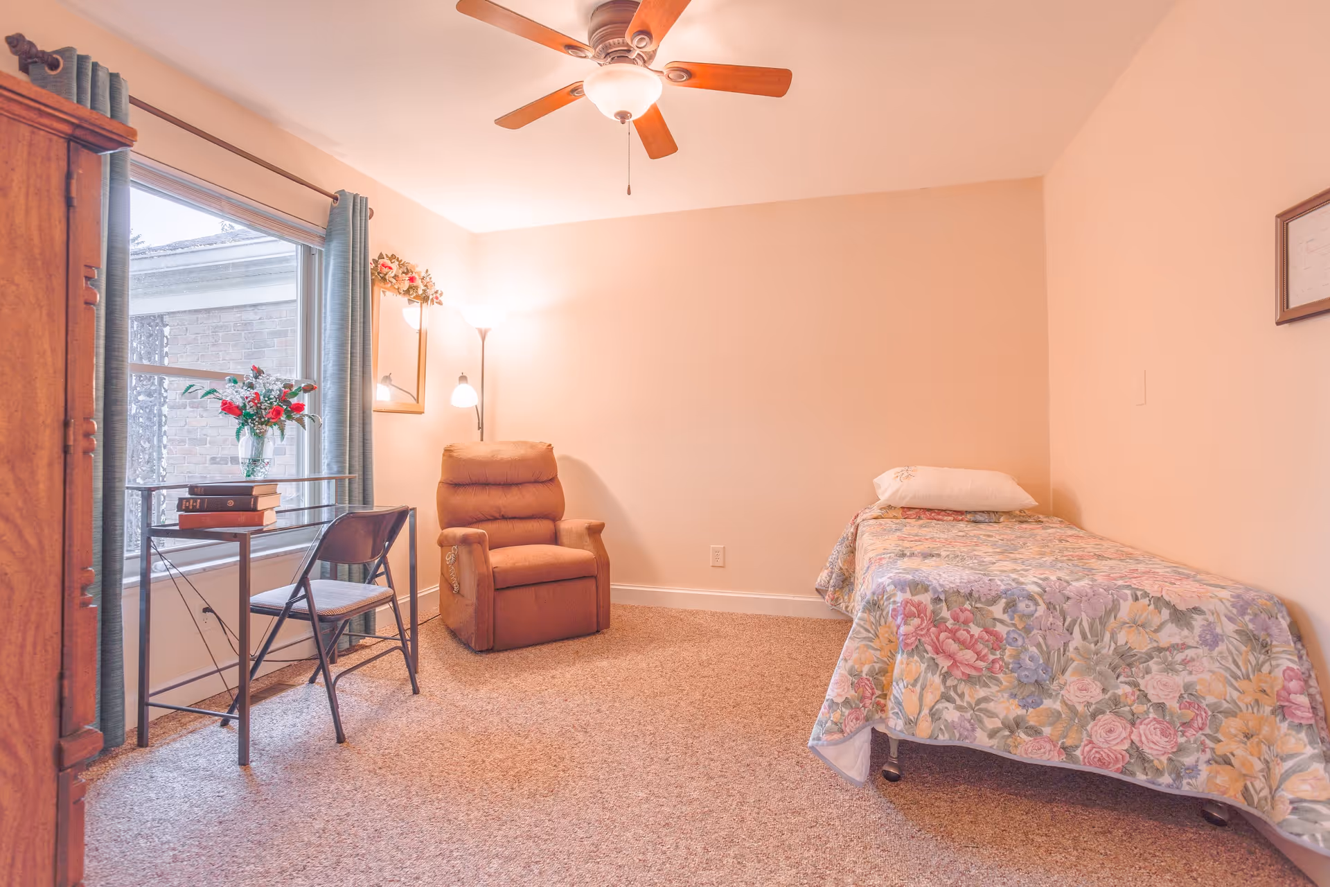 A cozy bedroom with a single bed covered in a floral bedspread, a brown recliner chair, a small desk with a chair, books, and a vase of flowers near a window with blue curtains. The room has beige walls, carpeted floor, a ceiling fan with light, and a floor lamp in the corner.