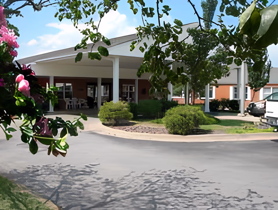Front exterior view of a senior living facility named Loch Haven, showing a covered entrance with white pillars, surrounded by green bushes and trees with pink flowers in the foreground under a partly cloudy sky.