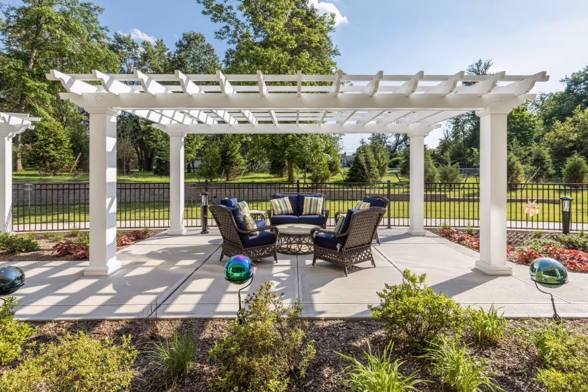 Covered outdoor patio with a white pergola over wicker seating and a table, surrounded by landscaping and a black fence.