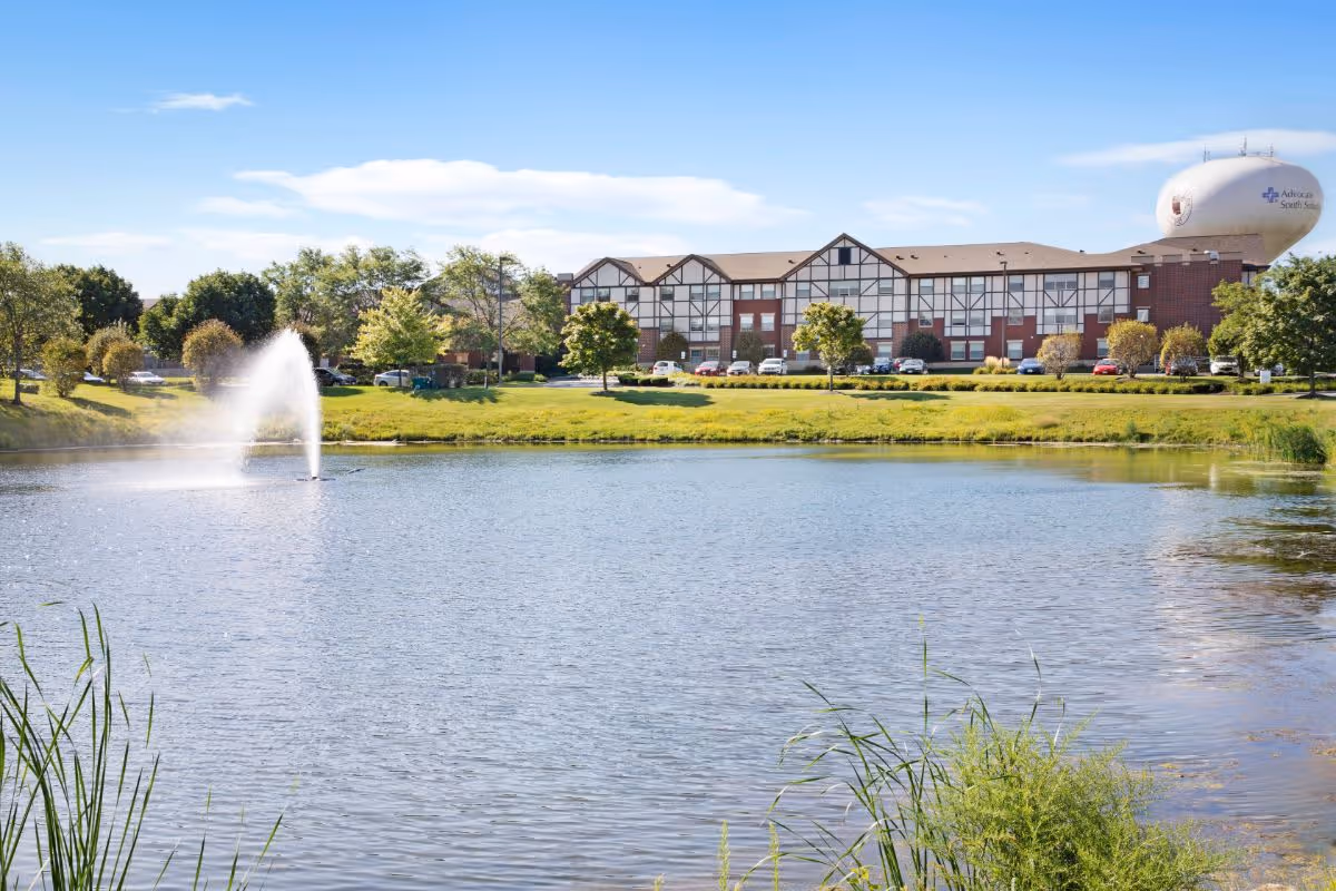 A large pond with a water fountain in the center, surrounded by green grass and trees. In the background, there is a multi-story building with a Tudor-style facade and a parking lot with several cars. A water tower with the text 'Advocate South Sub' is visible to the right of the building under a clear blue sky.