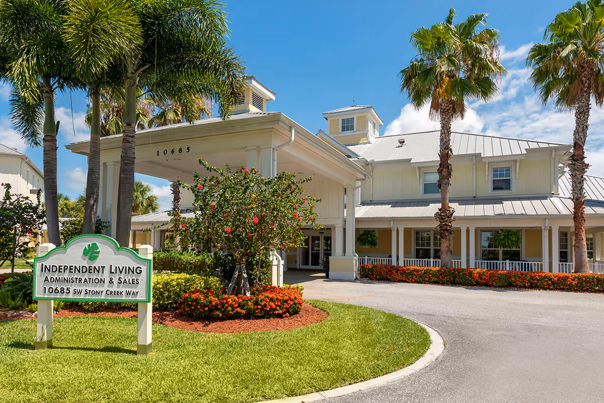 Front entrance of a yellow senior living building with palm trees, a driveway, and an 'Independent Living' sign on a landscaped lawn.