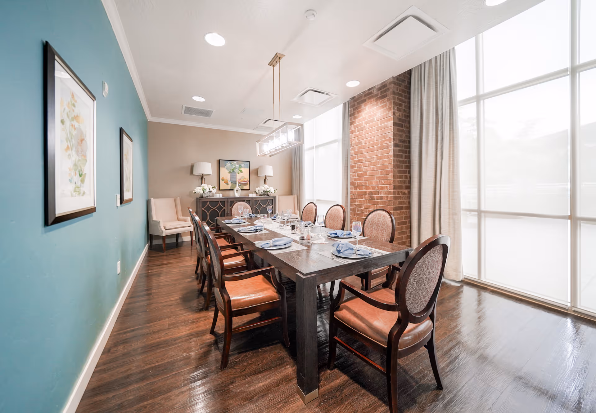 A well-lit formal dining room with a long wooden table set for a meal, upholstered chairs, large windows, and decorative wall art.