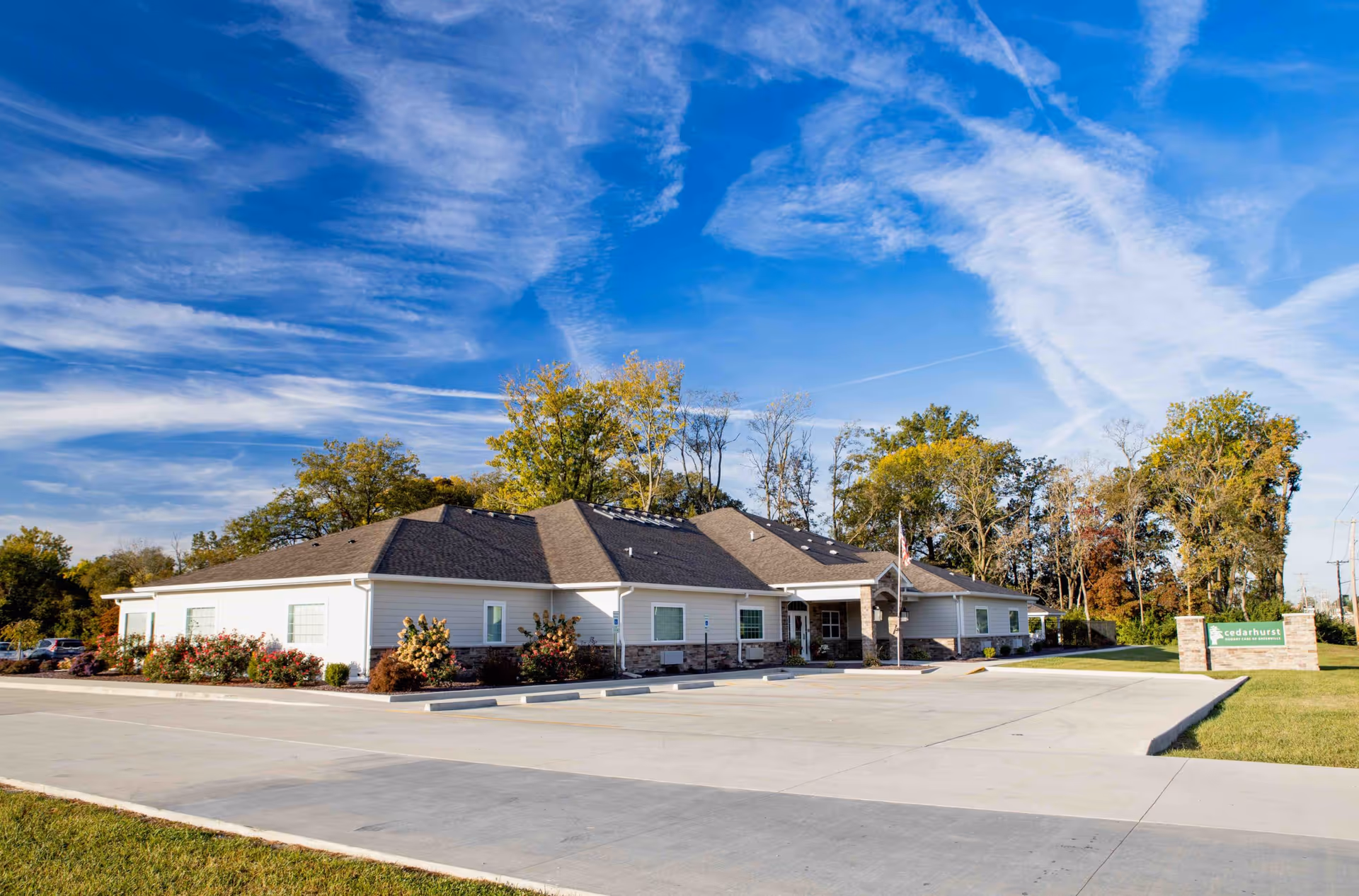 Exterior view of a single-story senior living facility building with a large parking area in front, surrounded by trees and a clear blue sky with some clouds.