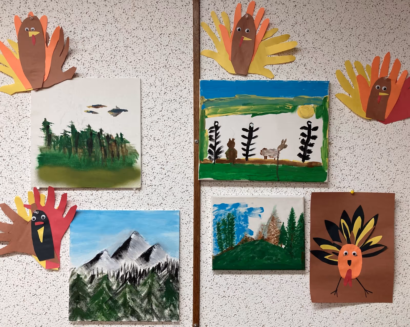A bulletin board displaying children's artwork including paintings of nature scenes with trees, mountains, and animals, surrounded by colorful paper hand turkeys made from cut-out hand shapes in autumn colors.