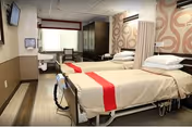 A rehabilitation and nursing center room with two hospital beds, each with white sheets and a red blanket at the foot. The room has a patterned accent wall, a window with blinds, a mounted TV, a sink, and a chair near the window. The floor is wood-style, and there is a privacy curtain between the beds.