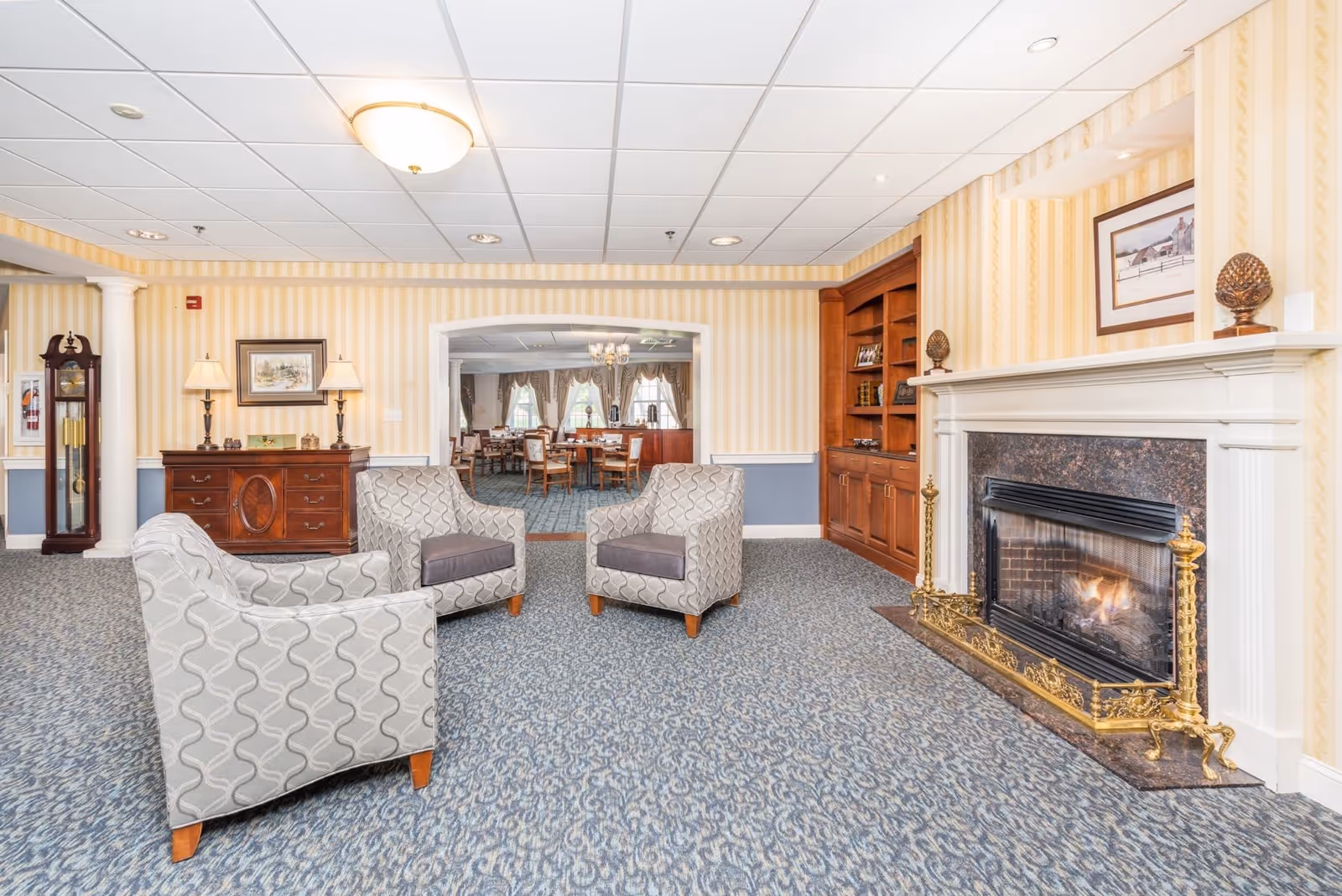 A cozy sitting area with three patterned armchairs arranged around a fireplace with a decorative brass screen. The room features a blue patterned carpet, yellow striped wallpaper, a wooden built-in bookshelf, and a wooden chest with two lamps and a framed picture above it. In the background, there is a dining room with tables and chairs, chandeliers, and large windows with curtains.