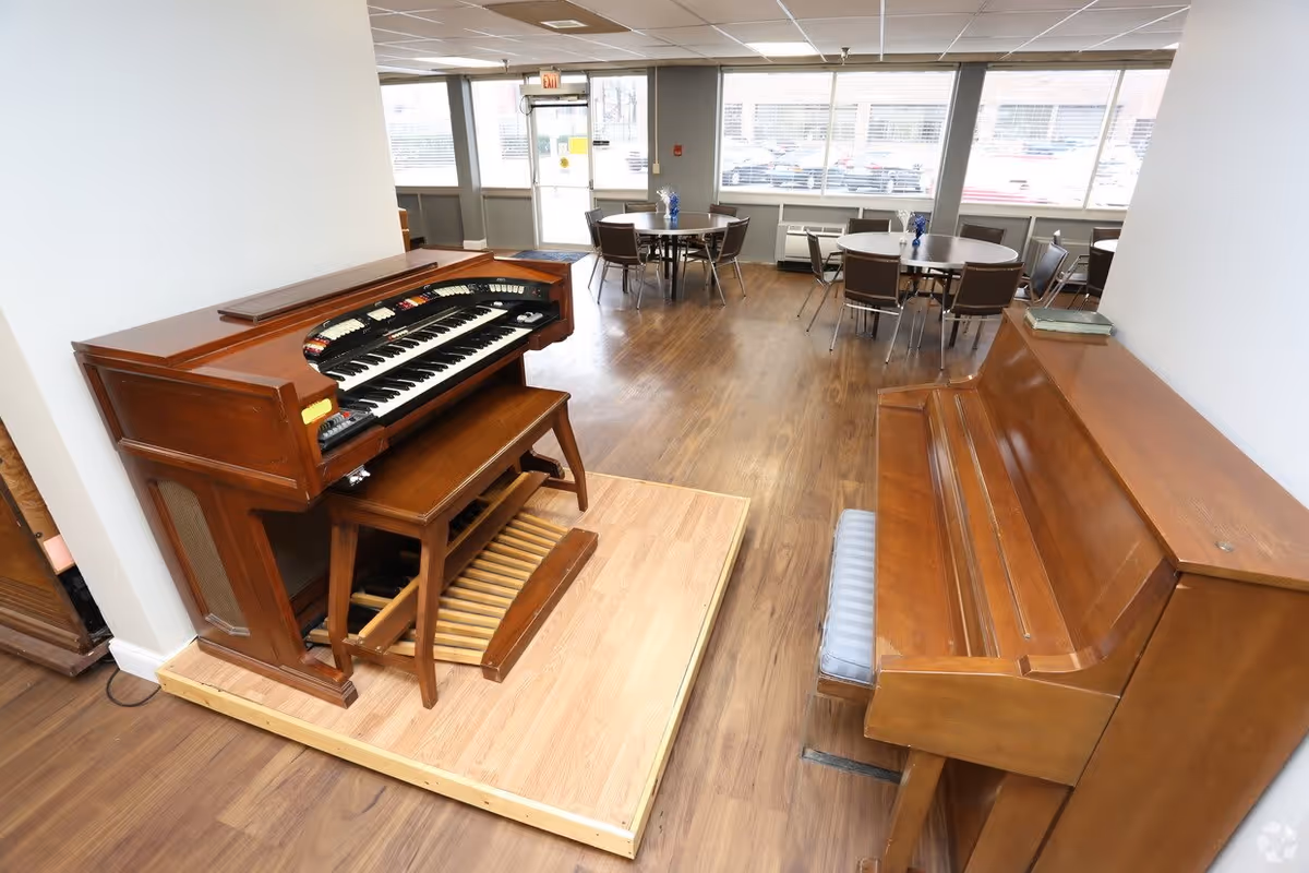 A spacious room with wooden flooring featuring a vintage organ on a small raised platform and a wooden upright piano with a cushioned bench. In the background, there are several round tables with chairs arranged near large windows letting in natural light.