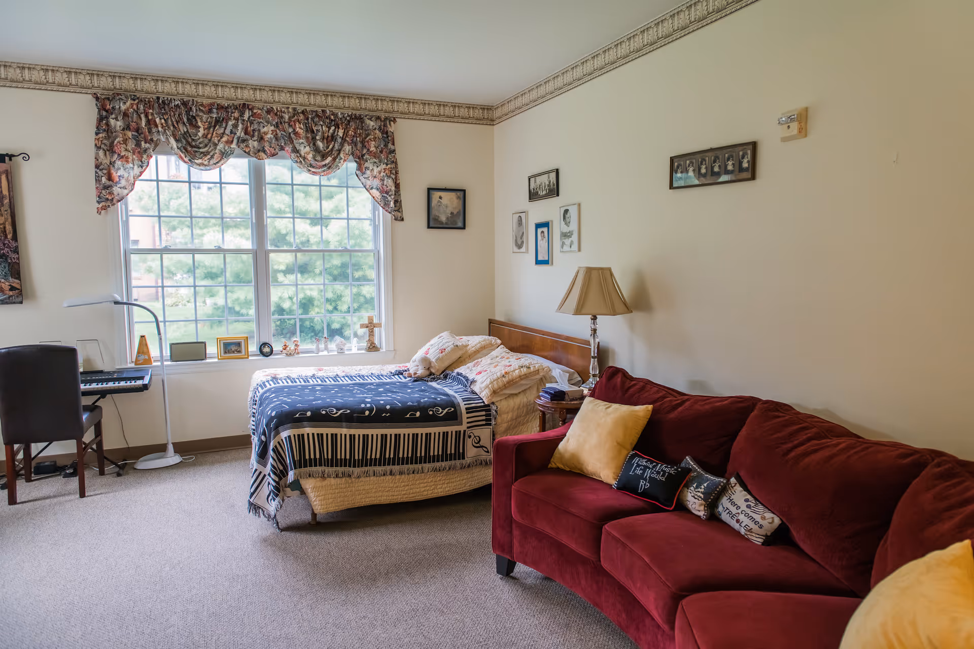 A cozy bedroom in Kingston Residence of Marion featuring a bed with a musical-themed blanket, a red couch with decorative pillows, a side table with a lamp, framed pictures on the wall, and a large window with floral curtains letting in natural light. There is also a keyboard and chair near the window.