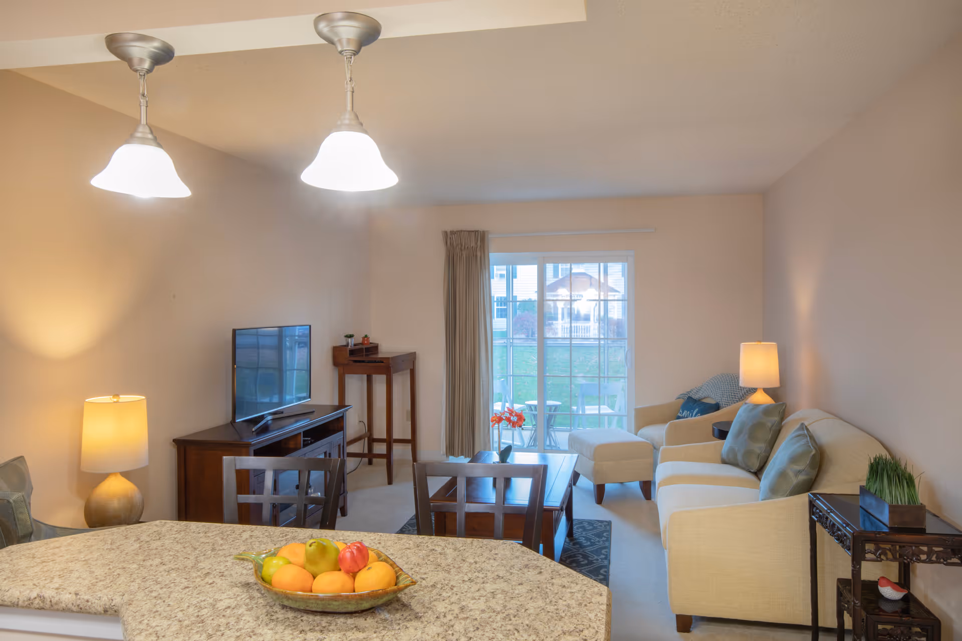 A cozy living room with a beige sofa, armchair, and ottoman, accented with green pillows. A wooden coffee table and side tables with lamps are present. A flat-screen TV sits on a wooden stand against the wall. Two pendant lights hang from the ceiling above a granite countertop with a bowl of fruit. Sliding glass doors lead to an outdoor patio with chairs and a table.