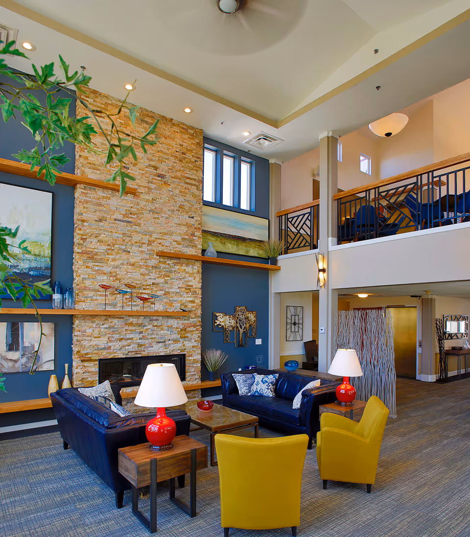 A spacious and modern living room area with high ceilings, featuring a stone fireplace wall with decorative shelves and artwork. The seating includes two dark blue leather sofas with patterned pillows, two yellow armchairs, and wooden side tables with red lamps. The room has a carpeted floor, large windows, and a balcony area with additional seating above.