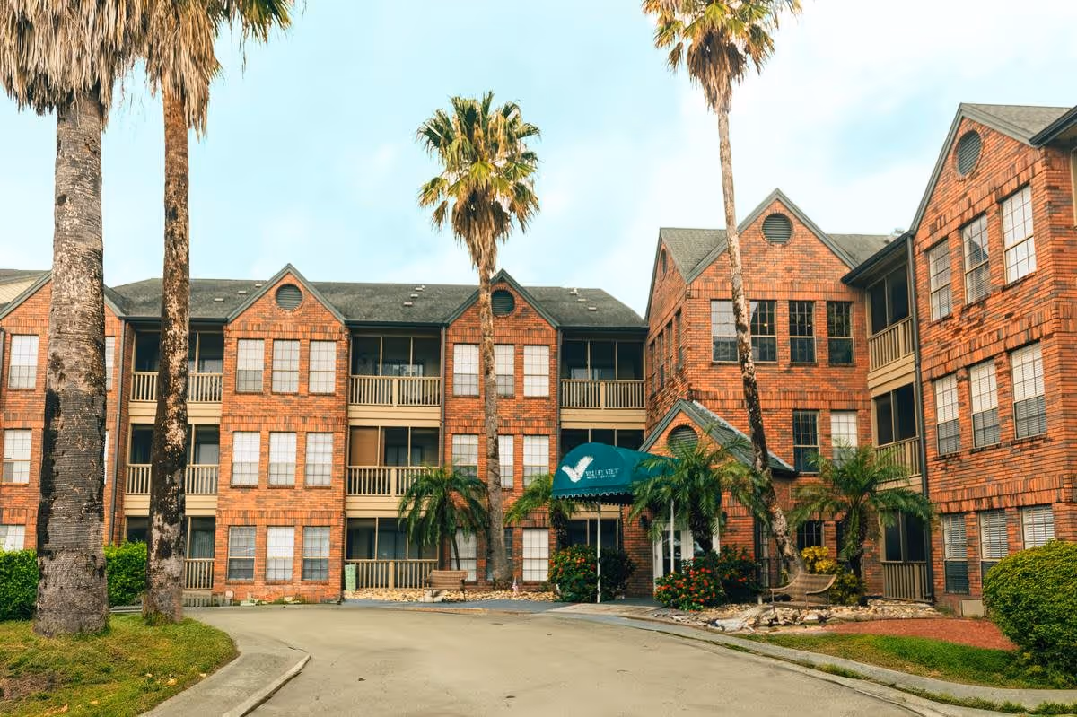 Exterior view of a three-story brick building with multiple windows and balconies, surrounded by palm trees and greenery. The entrance features a green awning with the name 'Valley View Senior Living' and a bird logo. There is a curved driveway leading to the entrance.