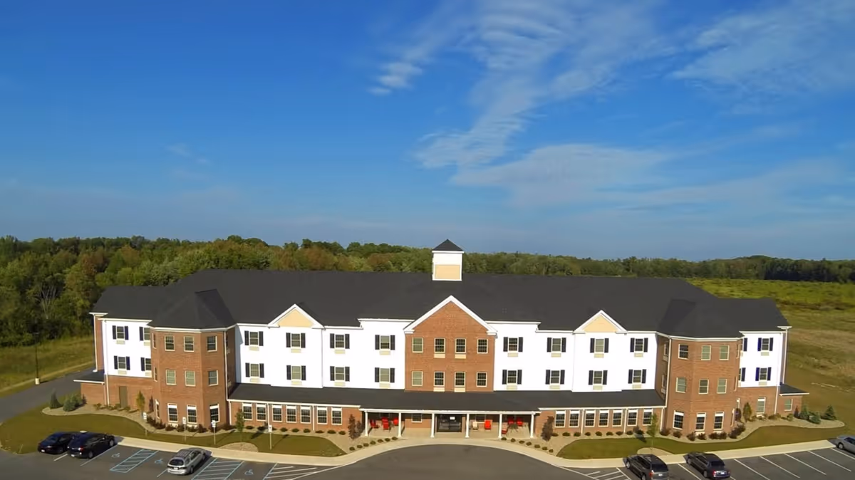 A large three-story assisted living facility building with a combination of brick and white siding exterior, surrounded by a parking lot with several cars and a backdrop of green trees and blue sky.