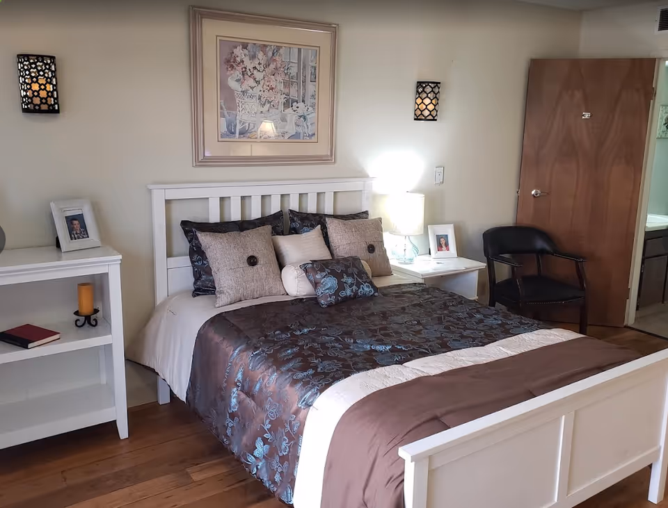 A neatly made bed with a white wooden frame and a brown and blue floral comforter. There are multiple pillows arranged on the bed. On the left side of the bed is a white shelf with a framed photo and a candle. On the right side is a white nightstand with a lamp and another framed photo. Above the bed hangs a framed floral painting. A wooden door and a black chair are visible to the right, with a glimpse of a bathroom in the background.