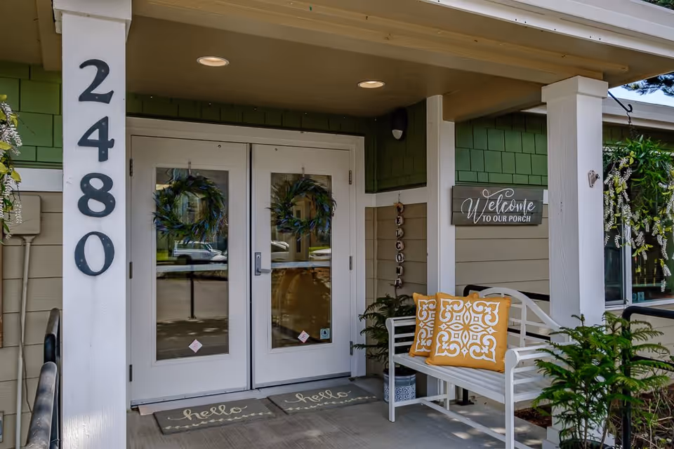 Covered entry porch showing double glass doors flanked by the address '2480', a bench with decorative yellow pillows, and a 'Welcome to Our Porch' sign.