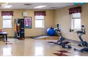 A spacious exercise room with two stationary bikes, a blue exercise ball, a rack of various exercise equipment, and a large mirror on the wall. The room has beige walls, two windows with maroon valances, and a polished floor with maroon diamond patterns.