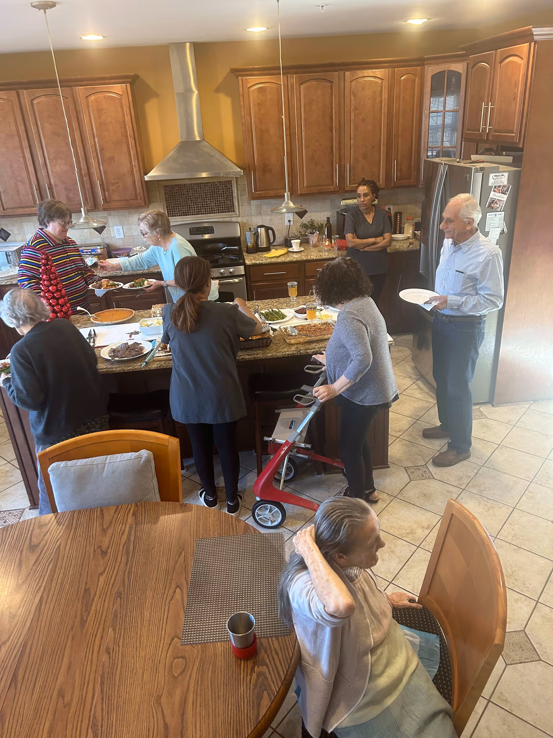 A group of older adults and caregivers gathered in a kitchen around a central island serving and eating a meal.
