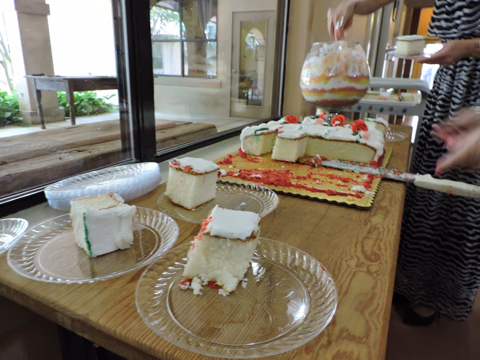 Slices of white frosted cake on clear plastic plates on a wooden table near a window. A partially cut cake with red and white frosting and decorative roses is on a gold-colored board. Two people are serving themselves dessert, one holding a glass bowl with layered dessert and the other holding a plate with cake.