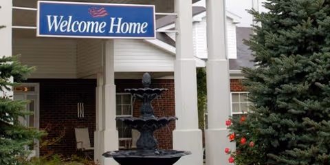 Entrance area of a residential facility with a 'Welcome Home' sign hanging above a black tiered water fountain. The building has white pillars and brick walls, surrounded by green pine trees and some red flowers.