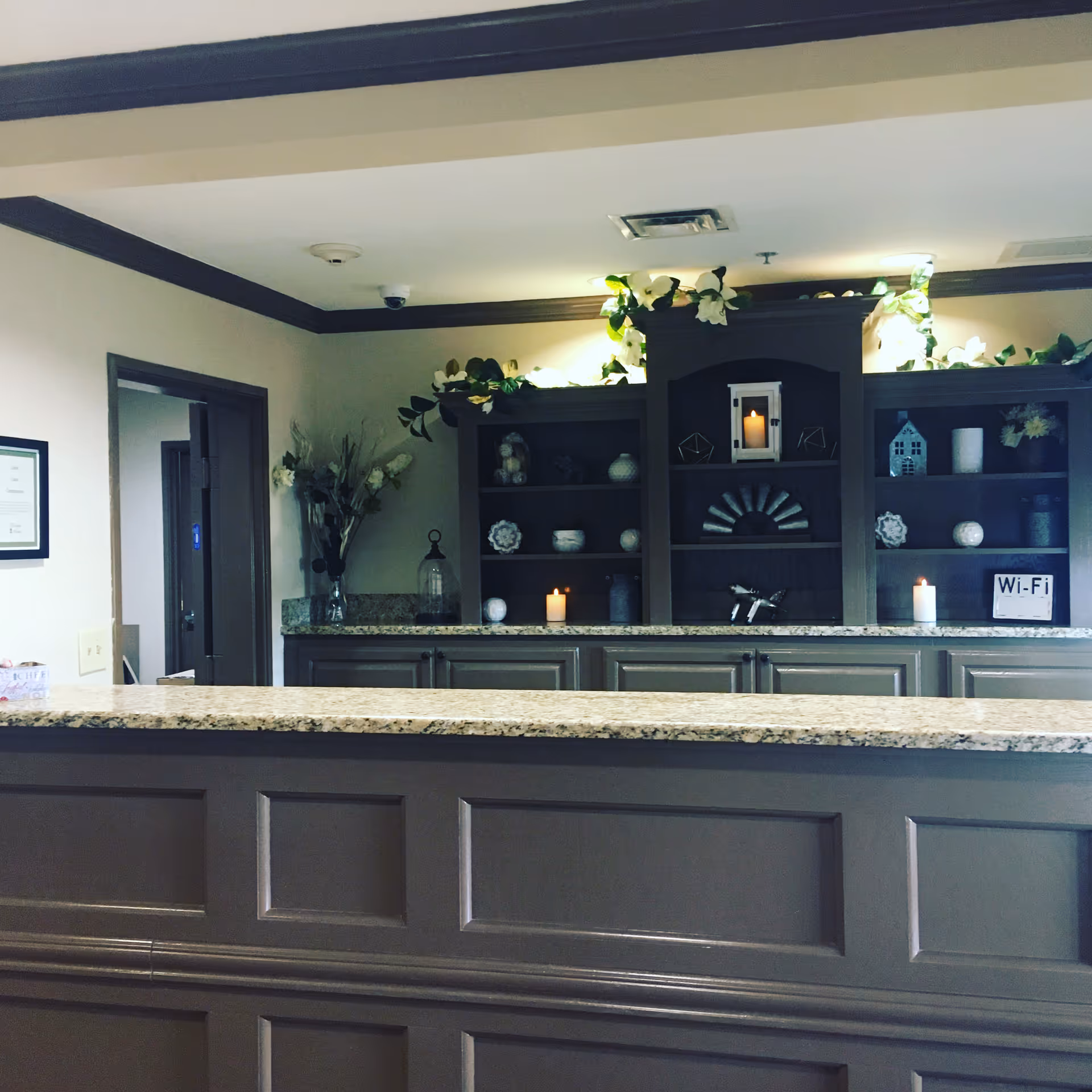 Reception area with a granite countertop desk in front of a dark wooden cabinet decorated with candles, small decorative items, and artificial flowers. There is a Wi-Fi sign on the cabinet and a doorway leading to another room on the left.