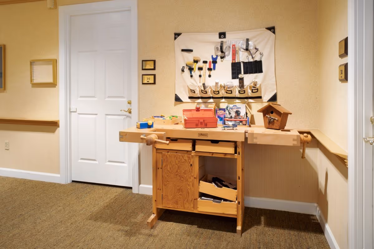 A wooden woodworking bench with tools, a small birdhouse and a wall-mounted tool organizer in a beige hallway interior.