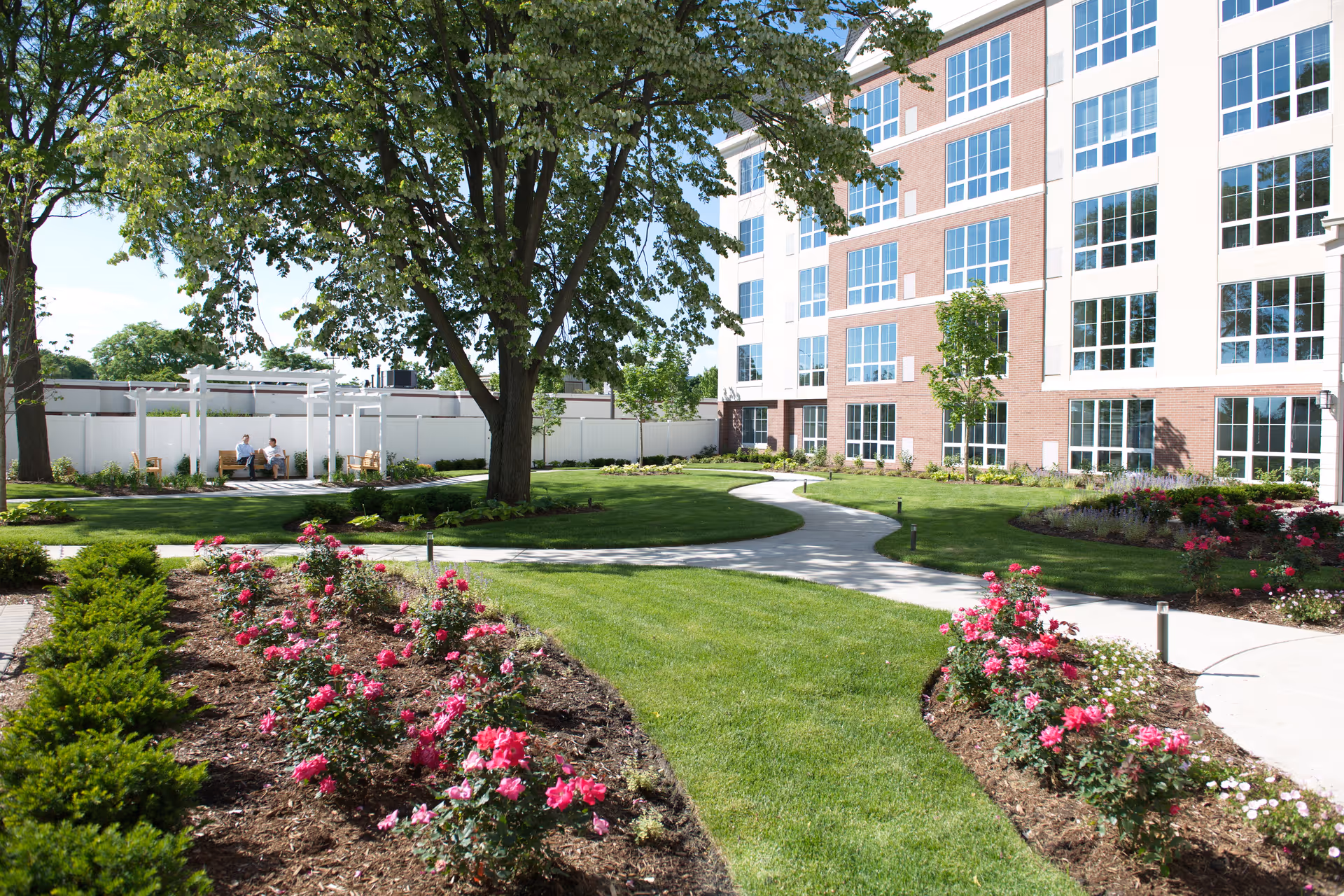 A landscaped outdoor garden area at the Grand at Twin Lakes facility featuring green grass, flower beds with pink flowers, a large tree, a winding concrete pathway, and a multi-story building with large windows in the background. Two people are seated on a bench under a white pergola.