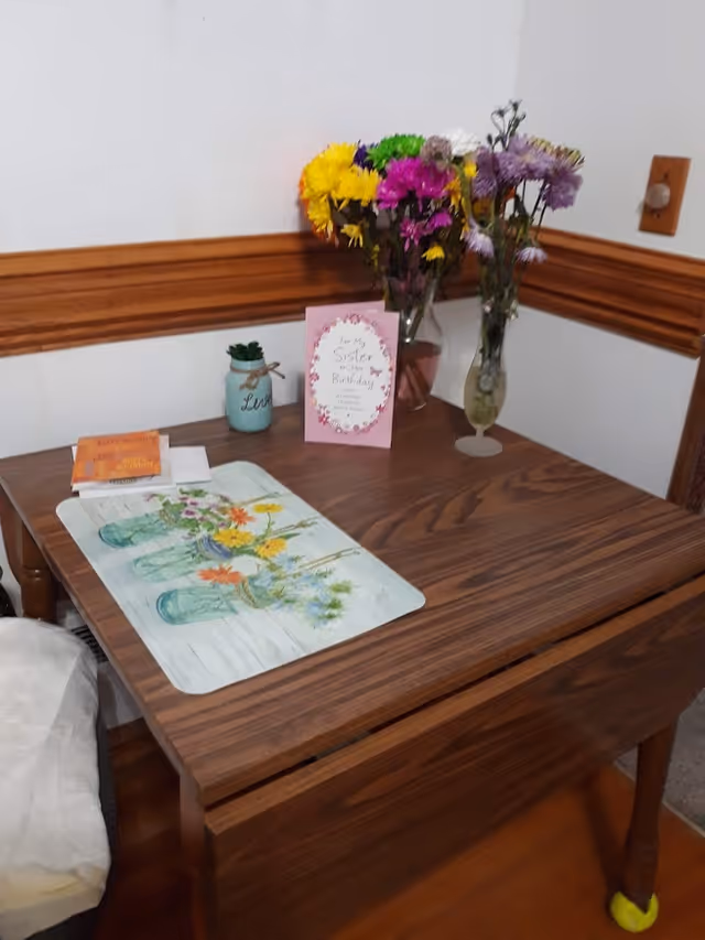 Wooden drop-leaf table in a corner with vases of colorful flowers, a birthday card, and a floral placemat.