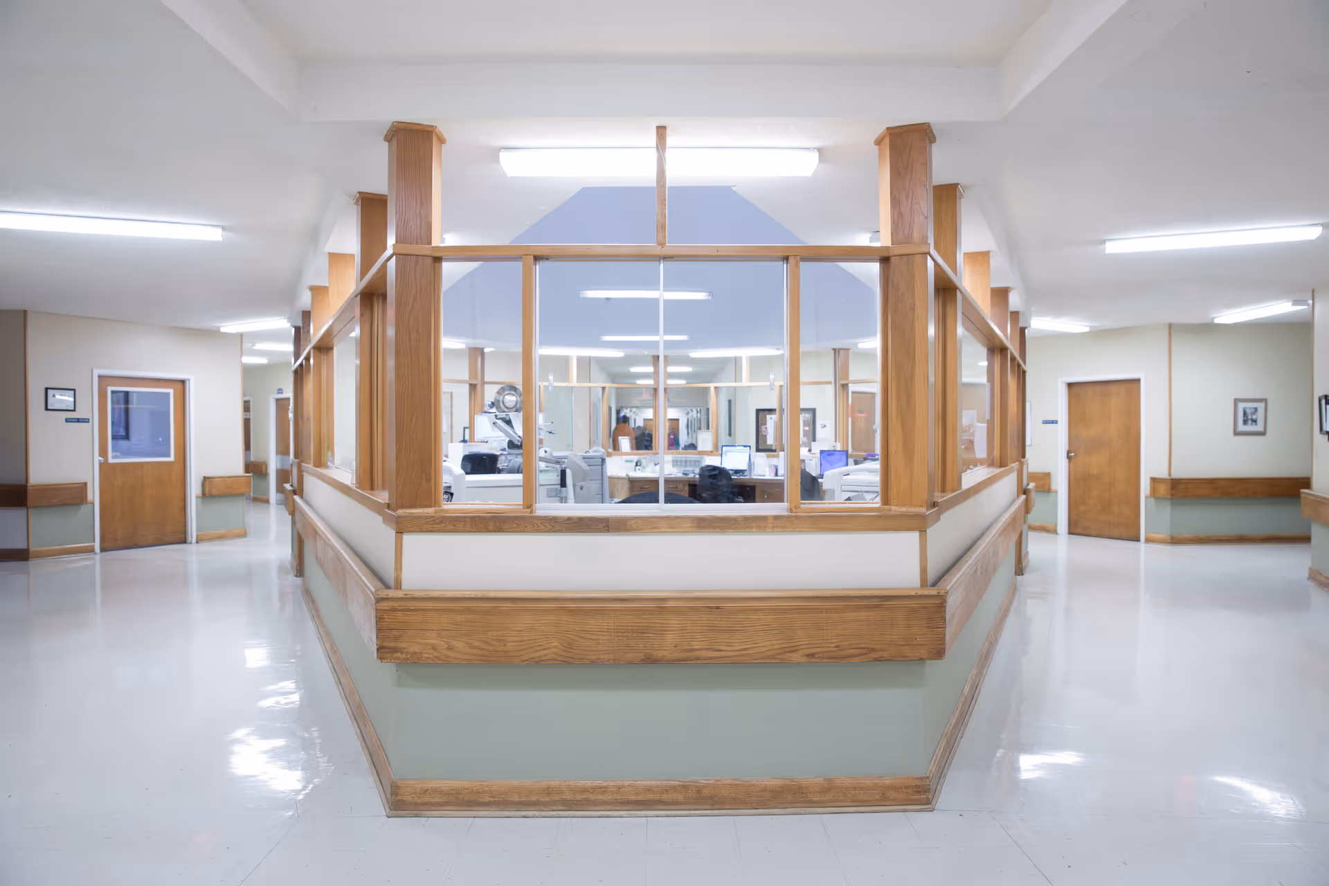 Interior view of a nursing station in a senior living facility with a central desk area enclosed by wooden framed glass panels. The surrounding hallway has doors and handrails along the walls, with bright overhead lighting and a clean, polished floor.