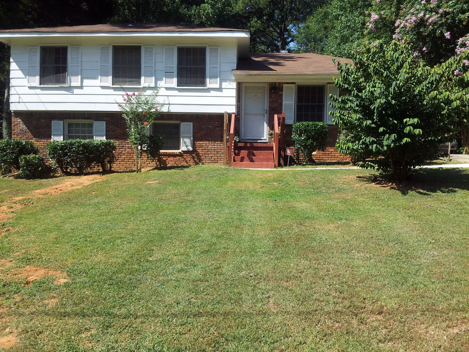 Front exterior view of a two-story house with white siding on the upper level and red brick on the lower level. The house has several windows with white shutters, a small set of red steps leading to a white front door, and a well-maintained lawn with bushes and a tree on the right side.