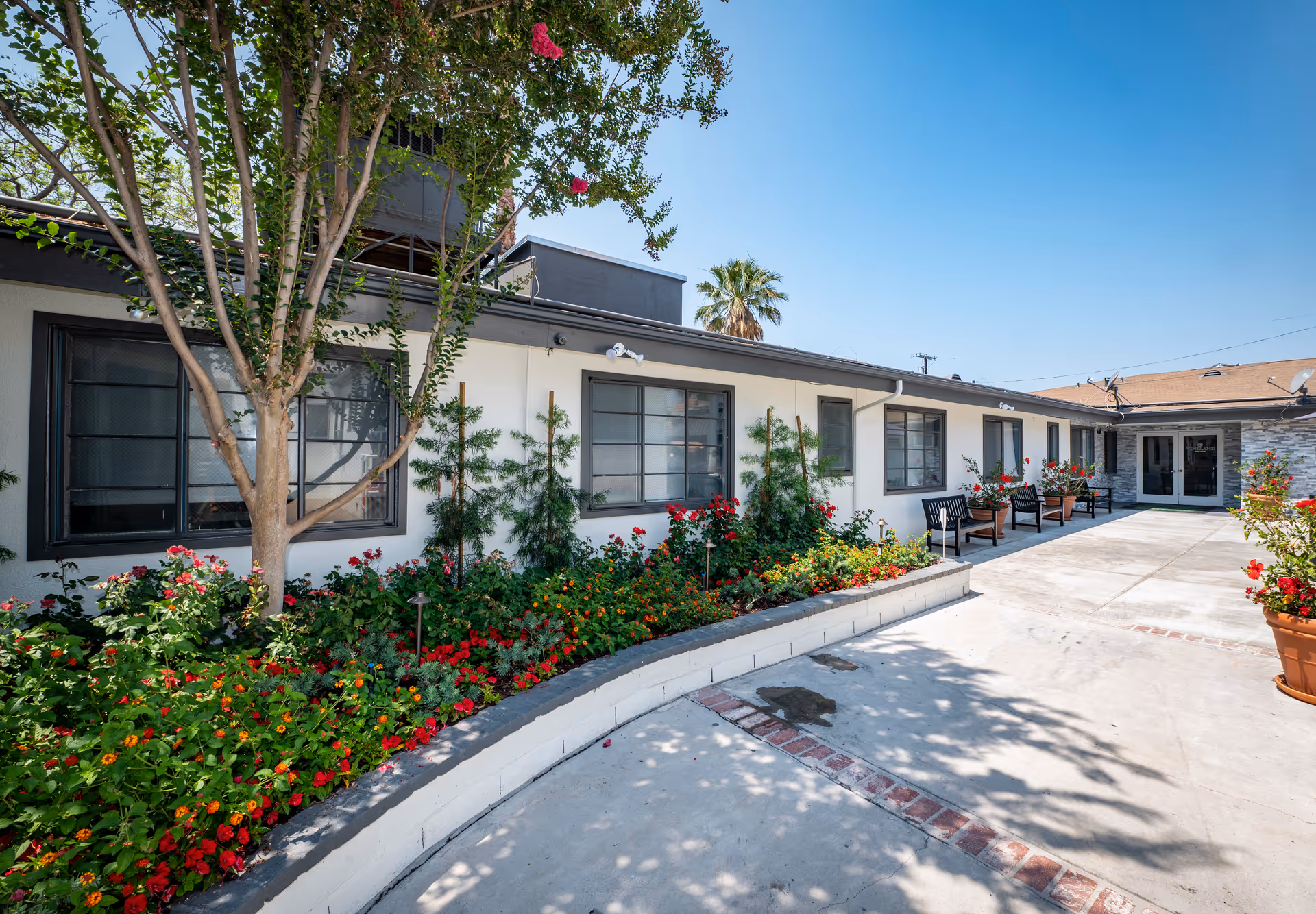 Exterior courtyard of a single-story senior living facility with flower beds, benches, and a paved walkway.