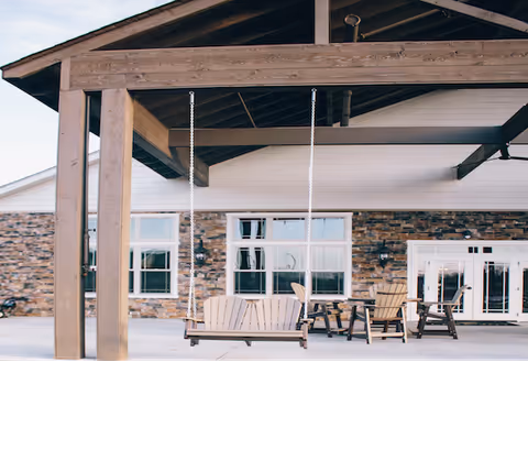 Outdoor seating area with a wooden swing hanging from a pergola structure, several wooden chairs and tables on a concrete patio, and a building with stone and white siding in the background.