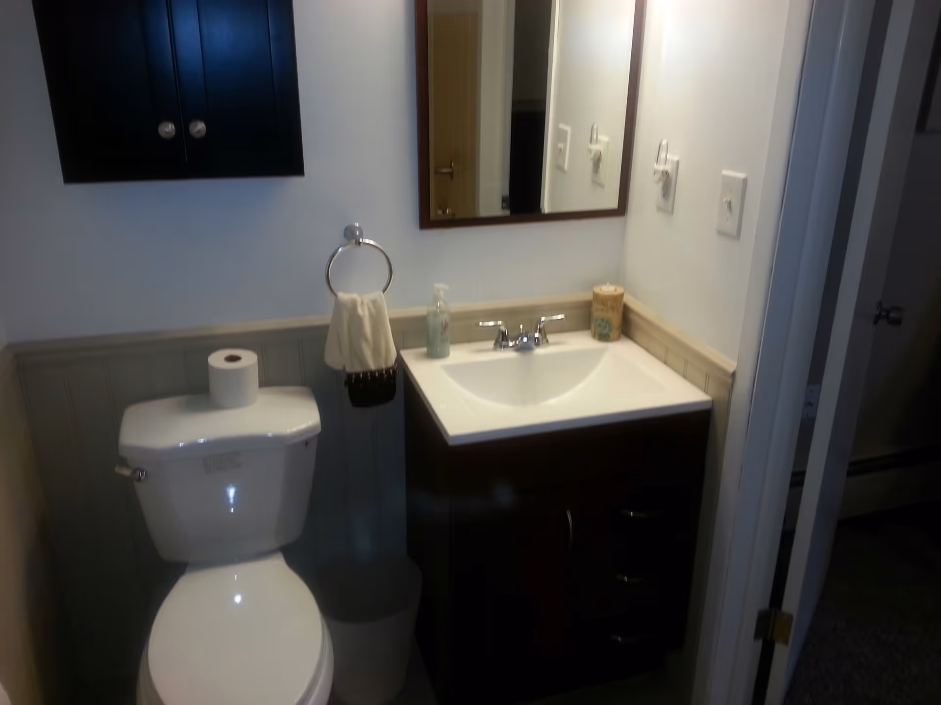 A small bathroom with a white toilet topped with a roll of toilet paper, a dark wood vanity with a white sink, a mirror above the sink, a towel ring holding a white hand towel, a soap dispenser, and a decorative container on the sink counter. A dark wood cabinet is mounted on the wall above the toilet.