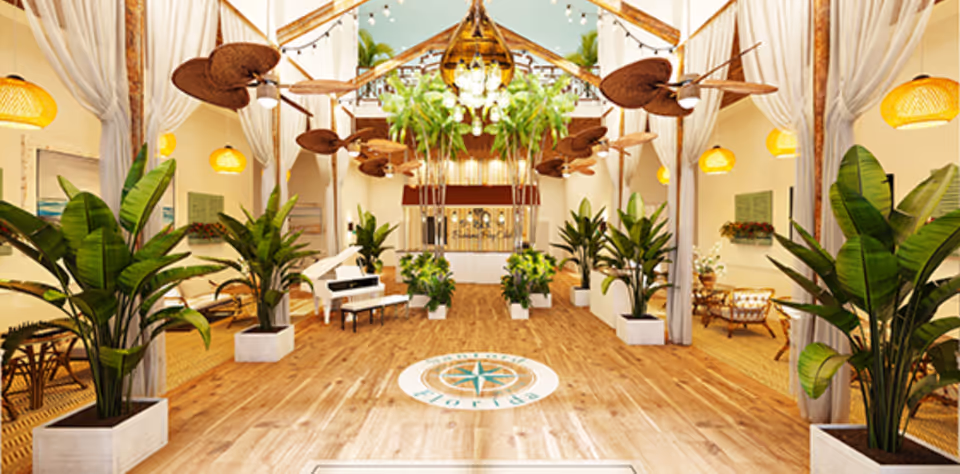 Spacious, well-lit interior of a senior living facility with wooden floors, large potted plants, ceiling fans with leaf-shaped blades, hanging yellow lamps, white curtains, and a white grand piano. The ceiling has exposed wooden beams and hanging greenery. The floor features a compass design with the text 'Bahama Bay Florida'.
