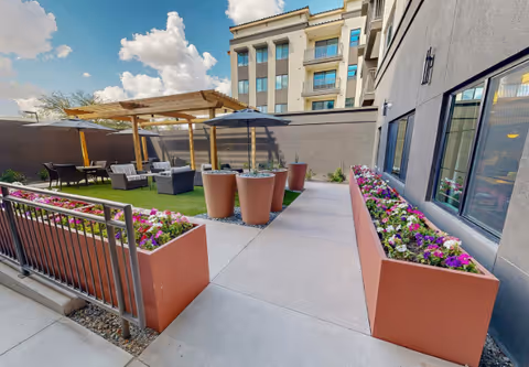 Outdoor patio area at Acoya Shea featuring a walkway flanked by large rectangular planters filled with colorful flowers. There are three tall round planters in the middle of the walkway. To the left, there is a seating area with cushioned chairs and a table under a wooden pergola, and an umbrella providing shade. The building exterior is visible on the right side with several windows, and a partly cloudy blue sky is seen above.