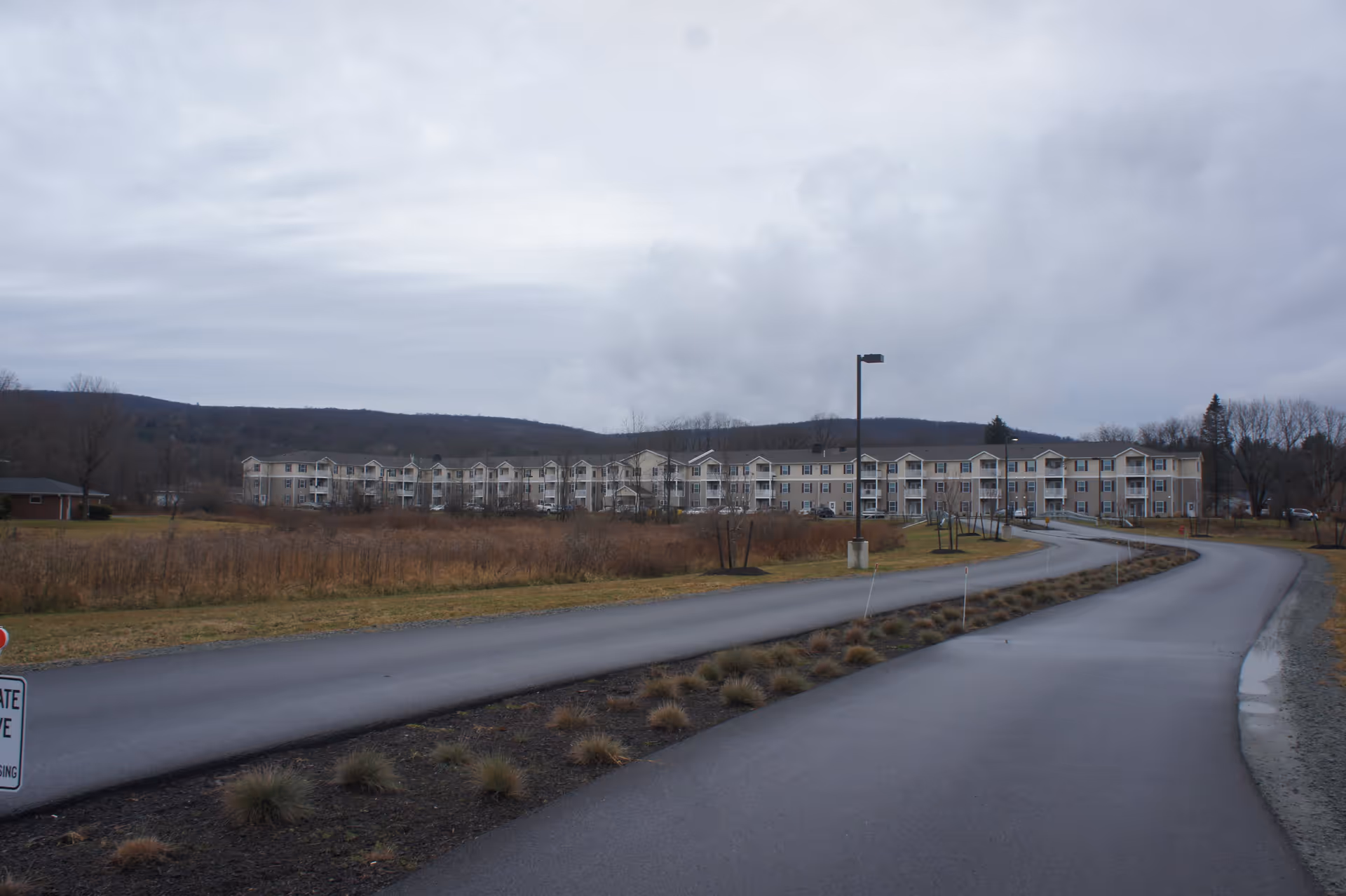 A long, curved paved driveway leading to a large, multi-story residential building in the distance, surrounded by open grassy fields and bare trees under a cloudy sky.