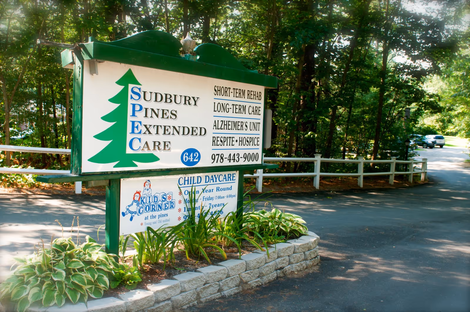 Outdoor view of a green and white sign for Sudbury Pines Extended Care, listing services such as short-term rehab, long-term care, Alzheimer's unit, respite, and hospice. The sign is surrounded by plants and trees, with a paved driveway and parked cars visible in the background.