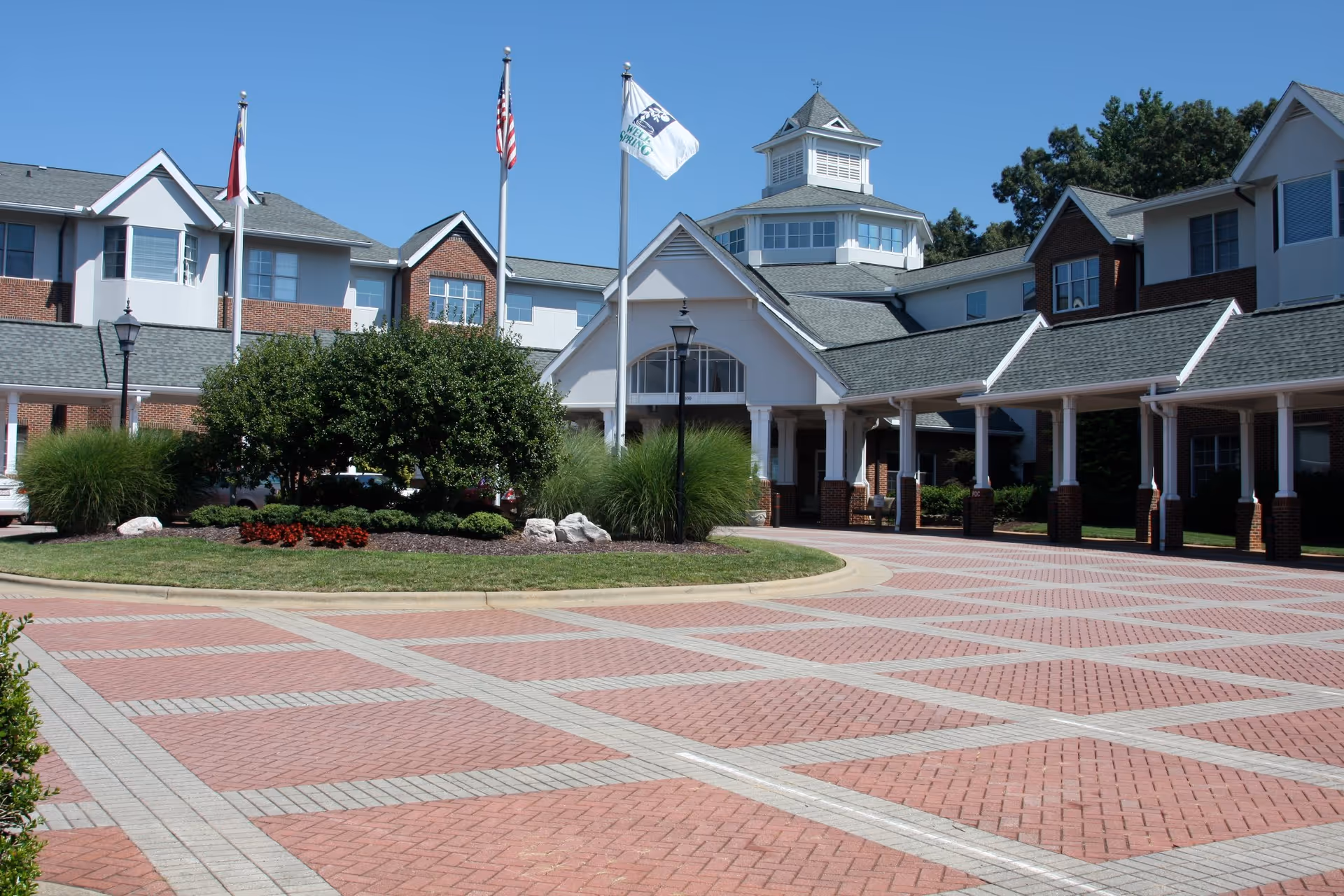 Front exterior view of Well-Spring Retirement Community building with a paved driveway, landscaped greenery, three flagpoles with flags, and a clear blue sky.