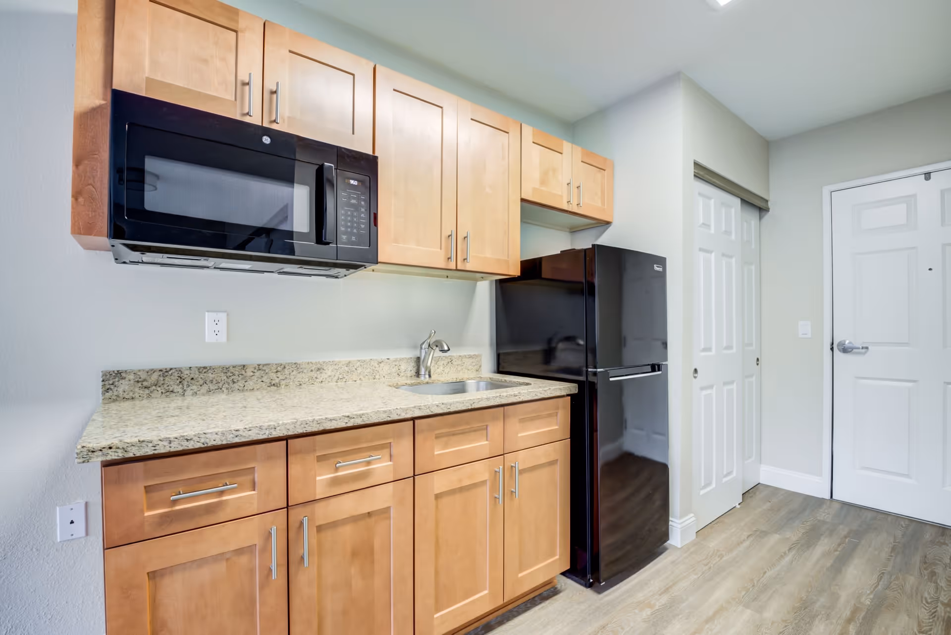 A small kitchen area with light wood cabinets, a granite countertop, a stainless steel sink, a black microwave mounted above the counter, and a black refrigerator next to a white door and a closet with sliding doors. The floor is light wood and the walls are painted light gray.