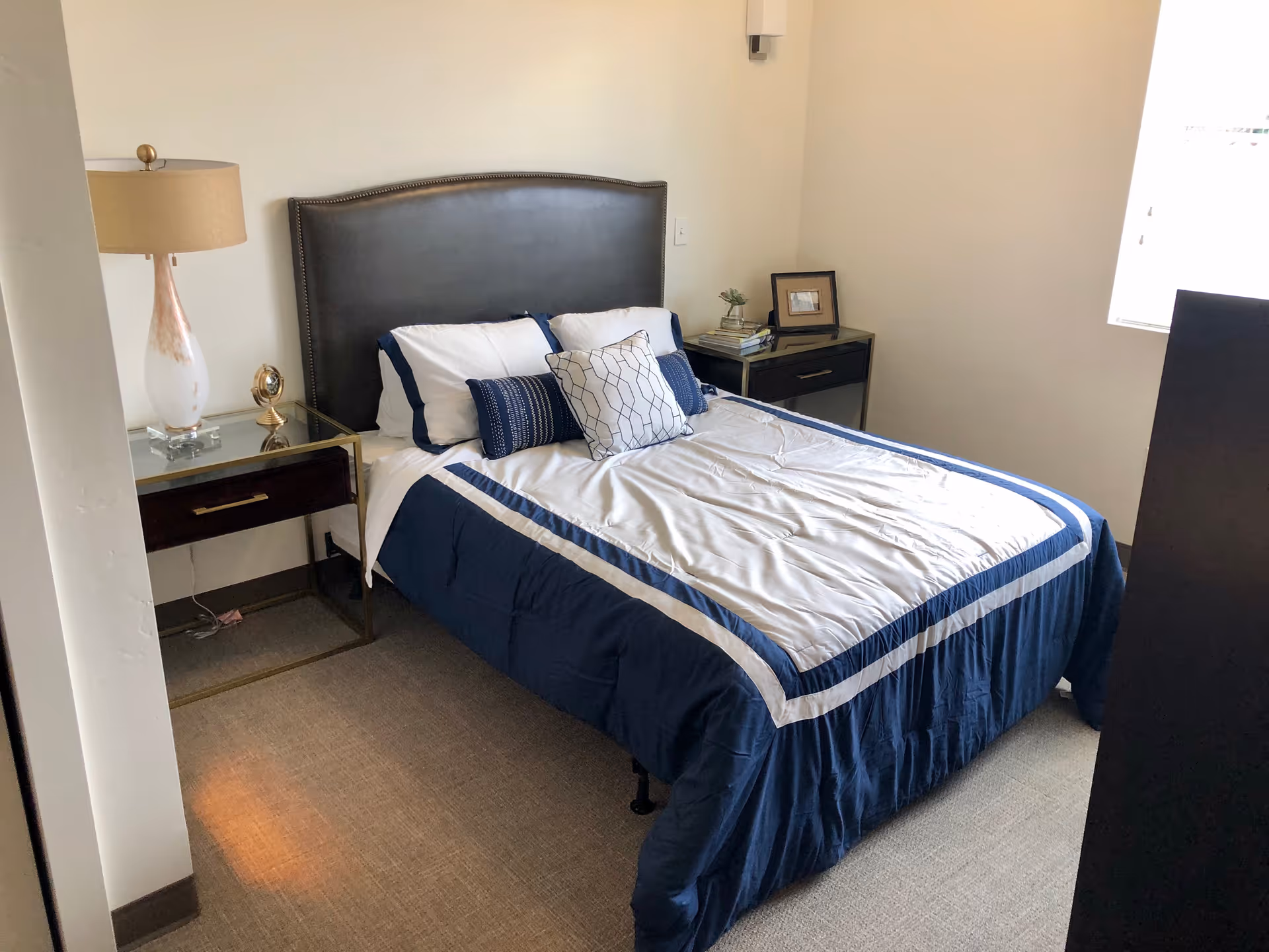 A neatly made bed with a dark upholstered headboard, white and navy blue bedding, and decorative pillows in a small bedroom. There are two nightstands on either side of the bed, one with a lamp and a small clock, and the other with books, a small plant, and a framed photo. A window on the right side lets in natural light.