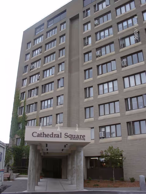 Exterior view of a multi-story senior living facility named Cathedral Square with a covered entrance supported by concrete pillars. The building has many windows, some with air conditioning units, and some greenery is visible around the entrance.