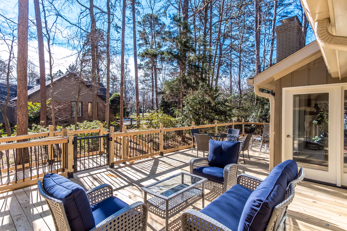 Sunlit wooden deck with blue-cushioned wicker seating and a glass-top table overlooking a wooded yard.