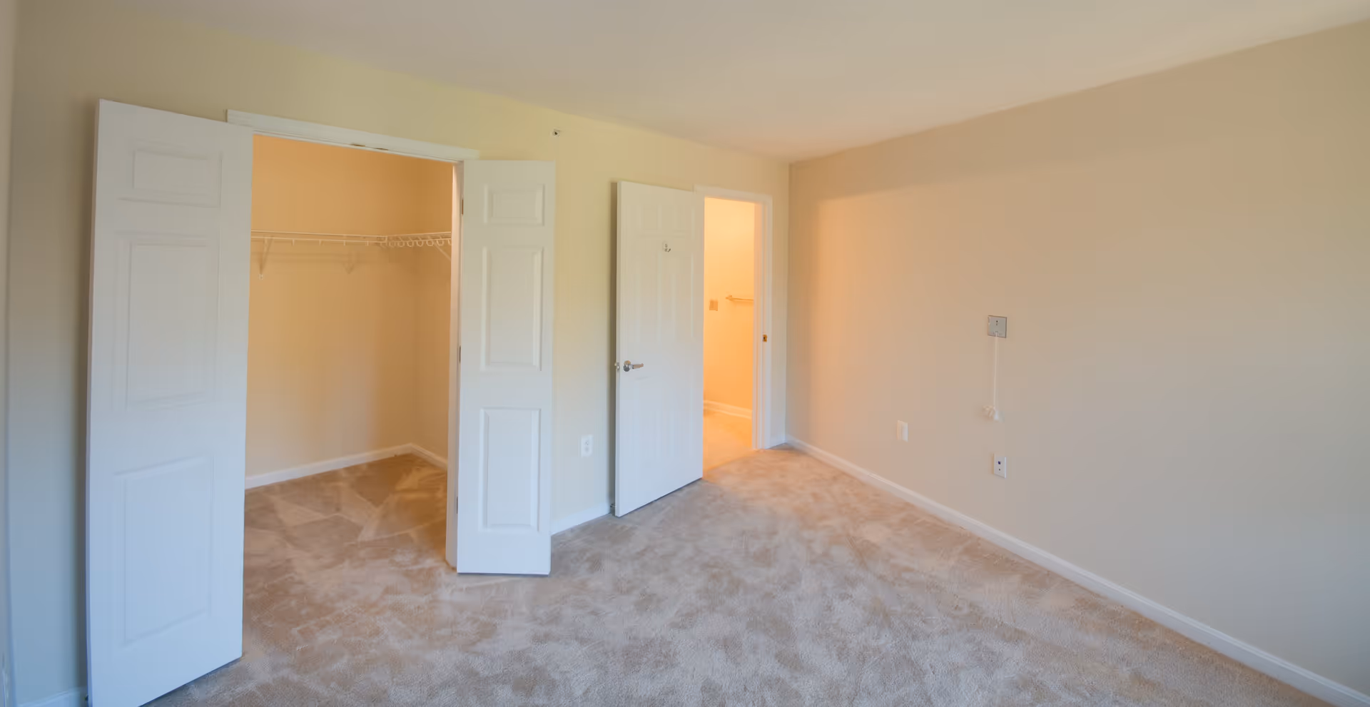 Empty bedroom with beige carpet and light yellow walls. The room features an open closet with white double doors and a door leading to another room with a tiled floor. There are electrical outlets and a wall plate on the right wall.