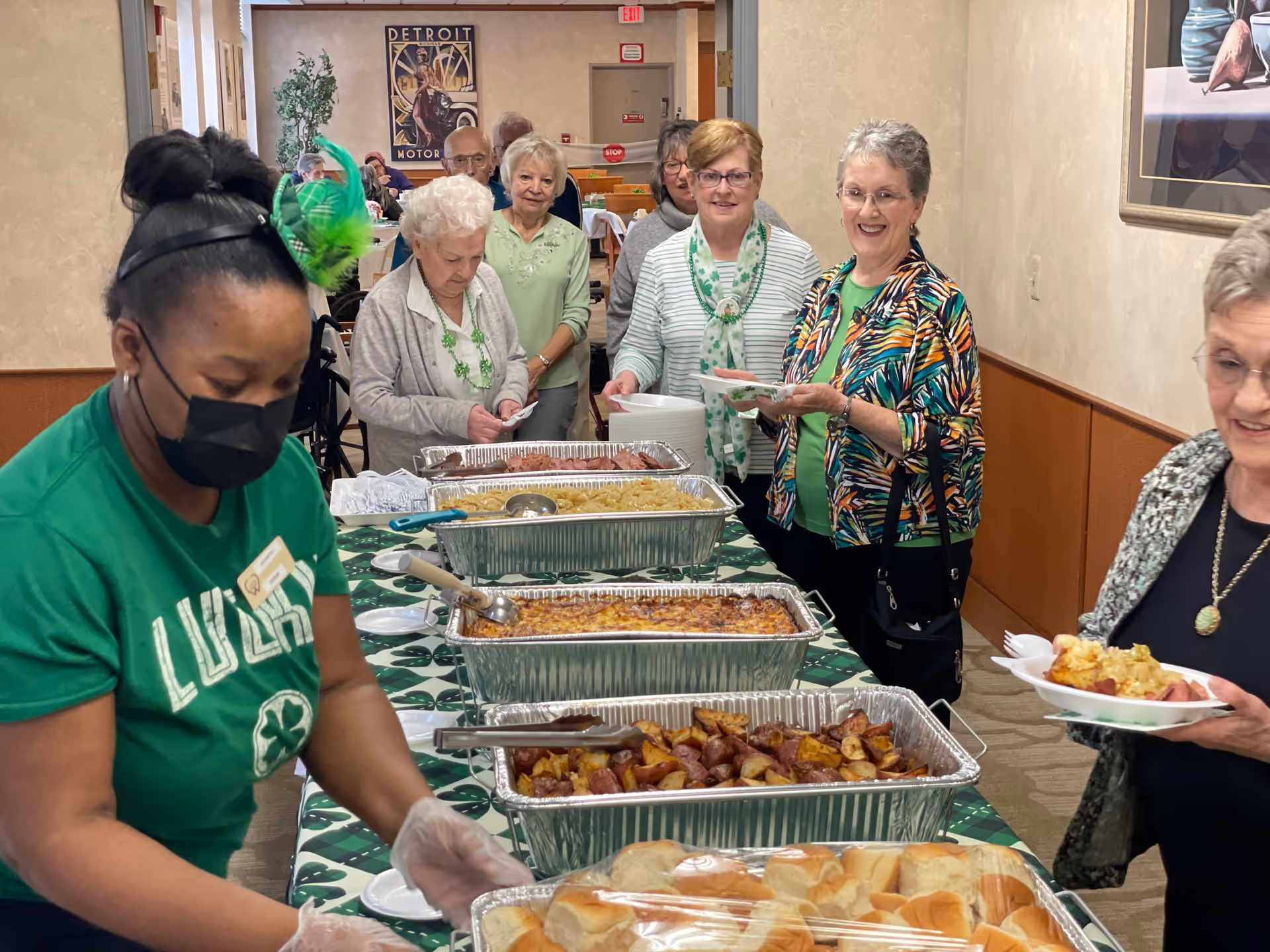 A group of elderly people standing in line at a buffet table with trays of food including rolls, roasted potatoes, pasta, and meat. A woman wearing a green shirt and a festive green headpiece is serving food. The setting appears to be a dining area with tables and chairs in the background.