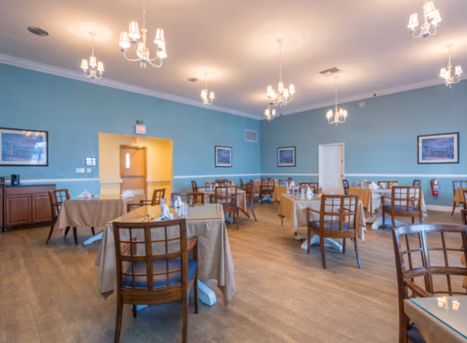 Dining room with several tables set with tablecloths and wooden chairs under chandeliers.