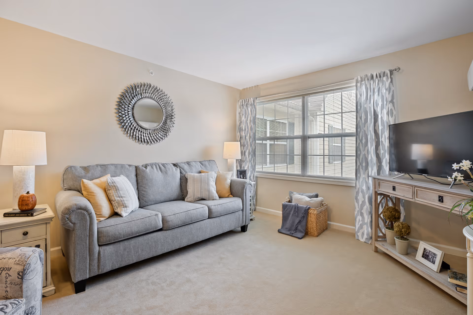 Bright furnished living room with a gray sofa, decorative round mirror, TV on a console, and a large window with patterned curtains.