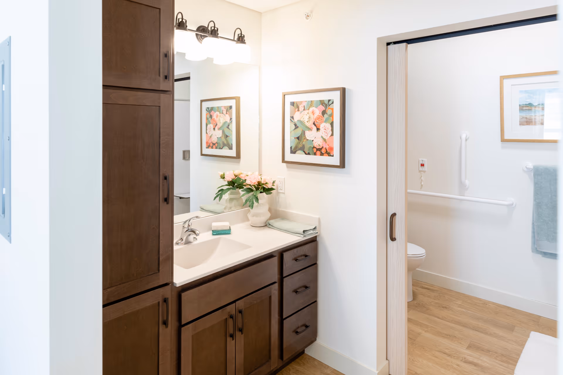 Well-lit bathroom vanity with dark wood cabinets, sink and mirror, and an adjacent toilet area with grab bars.