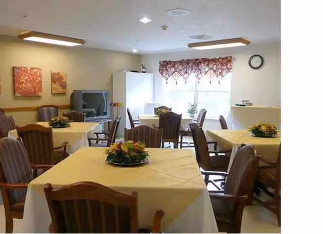 A dining room with several tables covered in yellow tablecloths, each decorated with a floral centerpiece. The room has wooden chairs with armrests, a window with a floral valance, a clock on the wall, and a television in the corner.
