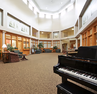 Spacious and well-lit common area in a senior living facility with high ceilings and large windows near the ceiling. The room features a grand piano in the foreground, multiple seating areas with green upholstered chairs and sofas, wooden framed windows and doors, potted plants, and framed artwork on the walls.