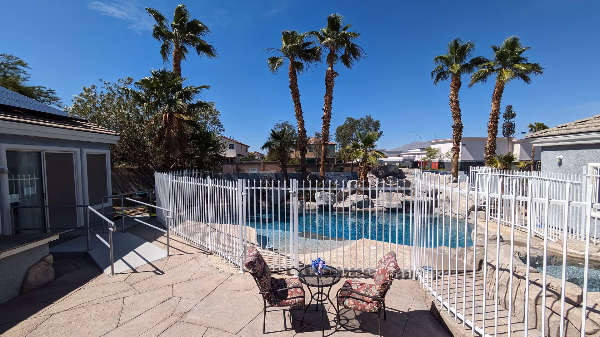 Fenced outdoor swimming pool and patio with two patterned chairs and a small table, surrounded by palm trees under a clear blue sky.