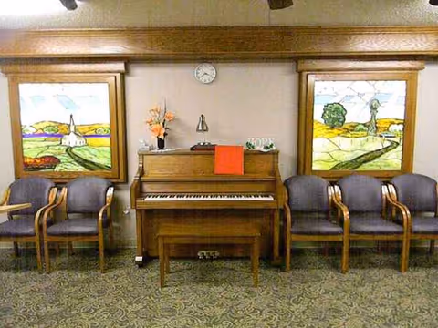 Center piano with orange cloth, flanked by chairs and colorful stained-glass panels.